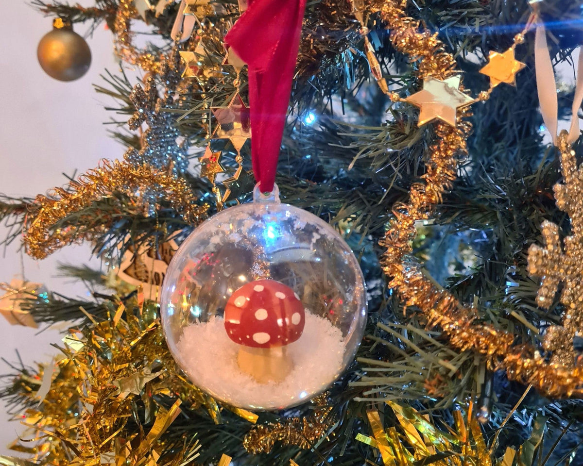 A handmade clear bauble snowglobe containing a handpainted wooden mushroom and snow, hanging on a Christmas tree with a red ribbon.