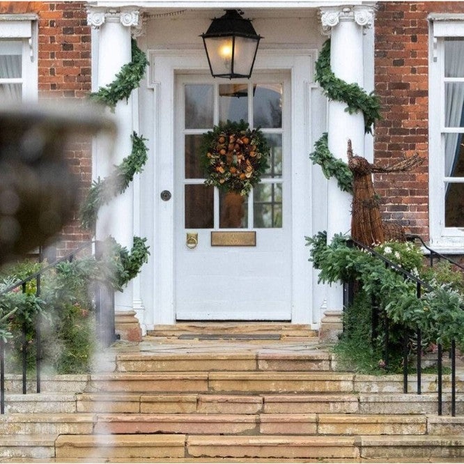 The white front door of Champneys, Henlow on a brick house porch decorated with green garlands, a floral wreath, stone steps, and a black lantern above the entrance.