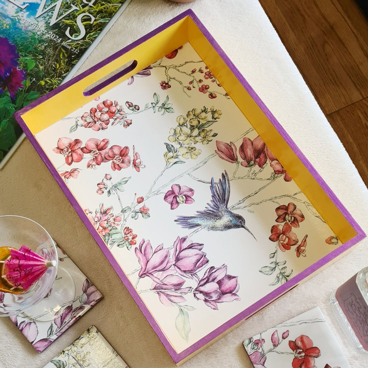 Angled view of handcrafted hummingbird floral serving tray with deep yellow painted sides and metallic violet edging.