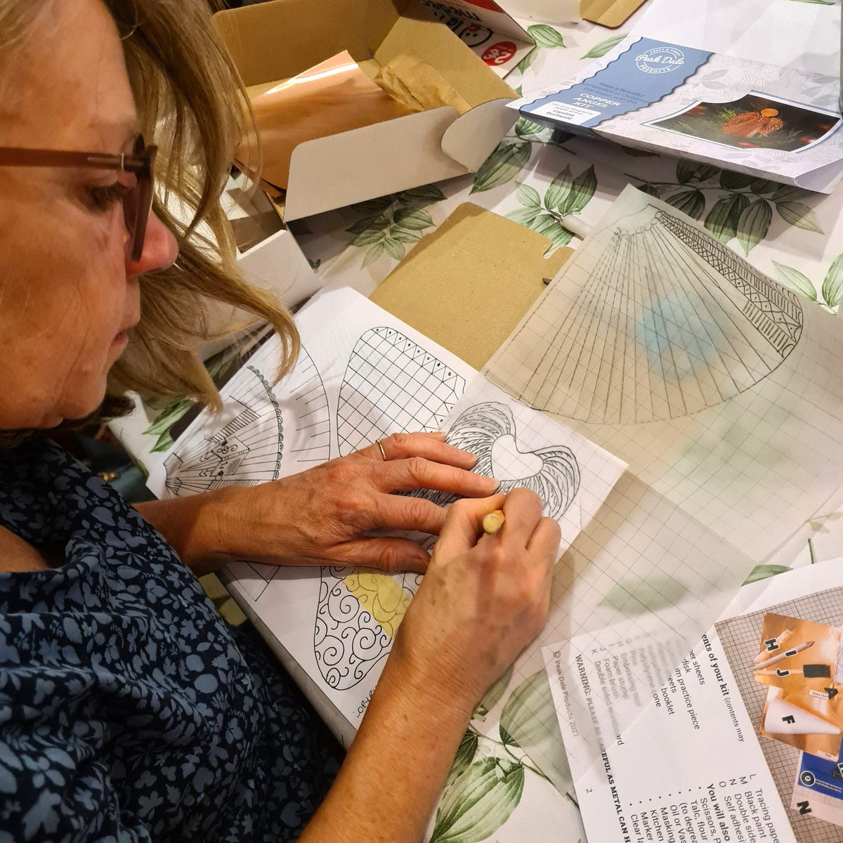 A woman with glasses is carefully making her template, ready to emboss part of her copper angel, surrounded by craft supplies, papers, and boxes.