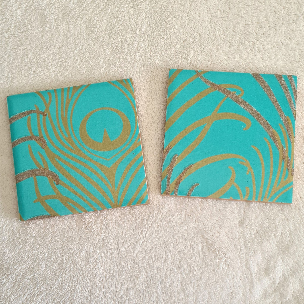 shimmering teal peacock feather design on a handmade ceramic coaster pair.