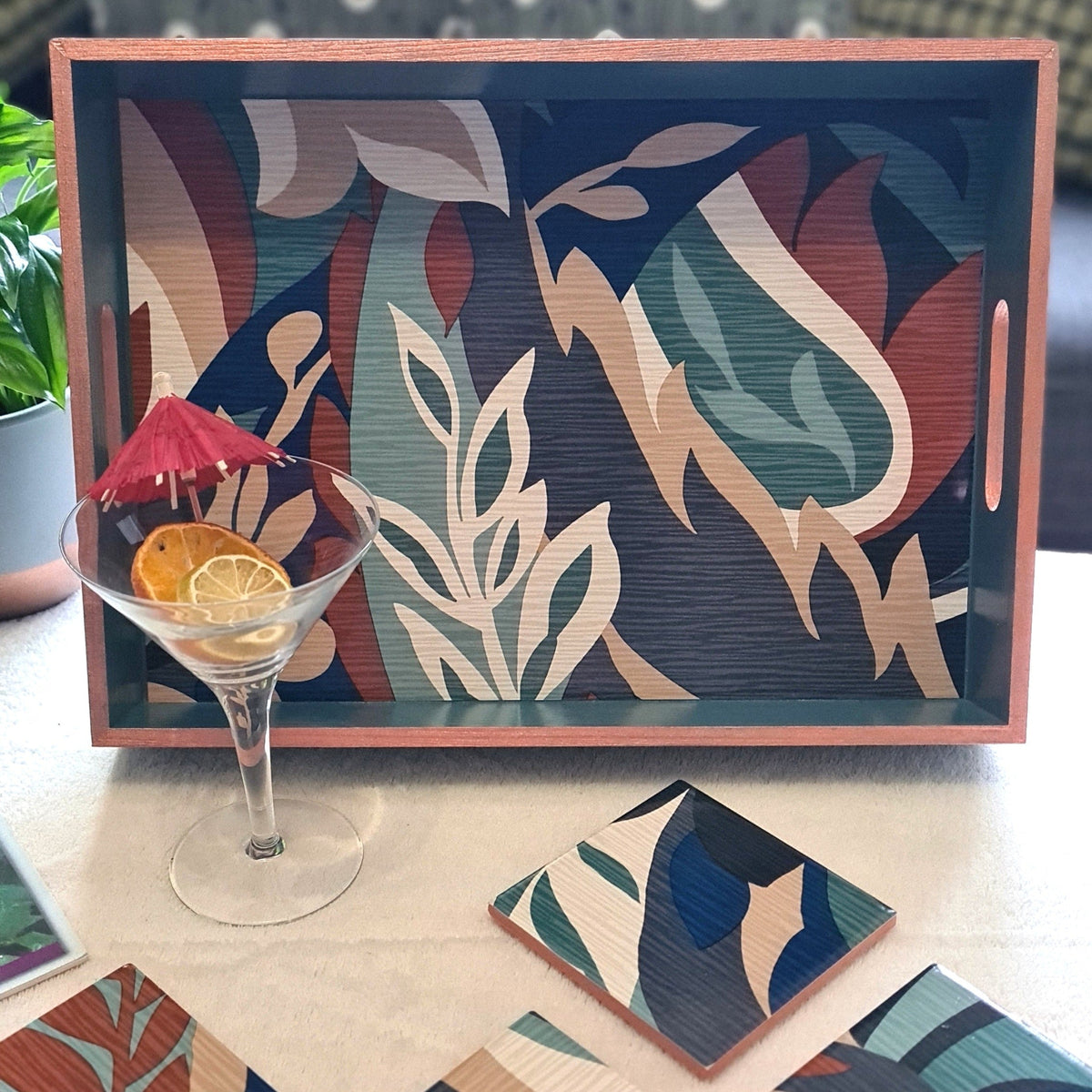 Handcrafted deep teal wooden serving tray decorated with a bold retro leaf design and metallic copper edging, styled on a coffee table with cocktail glass.