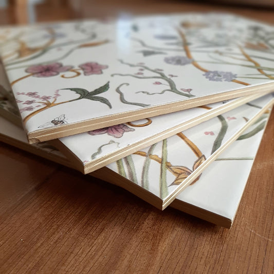 A stacked set of handmade ceramic table place mats with gold edges, created from Chateau Potagerie wallpaper, arranged on a wooden dining table.